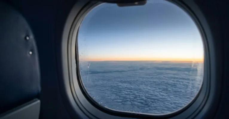 Janela de avião ao amanhecer, com céu azul e camada de nuvens abaixo